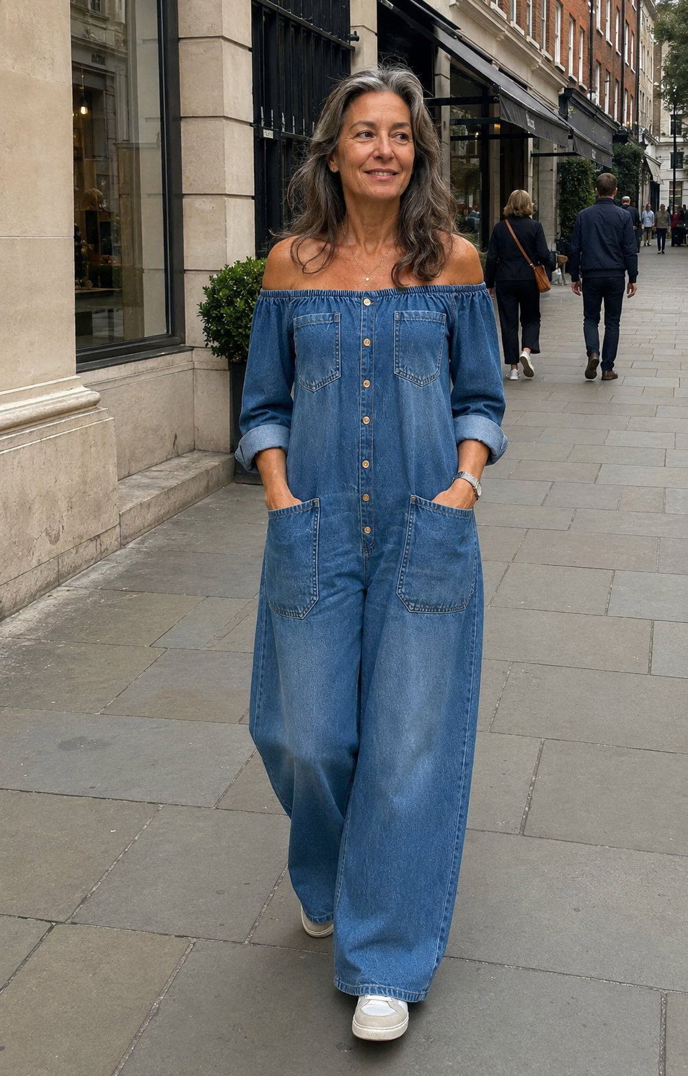 55-year-old woman walking in London wearing the blue Zaria off-shoulder denim jumpsuit